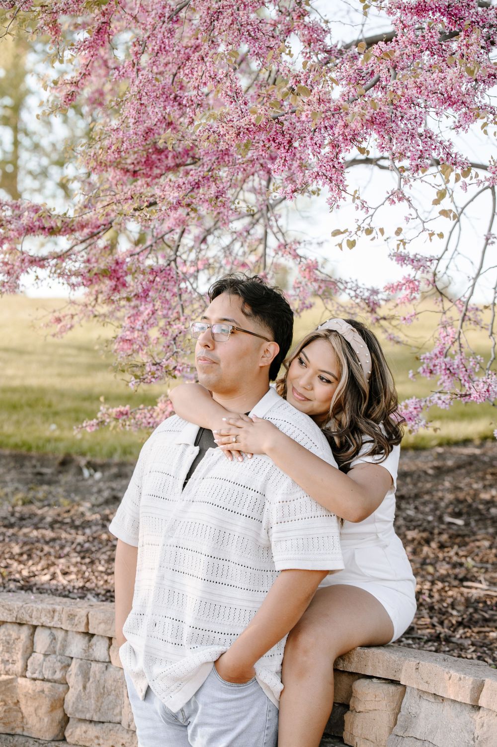 A couple shares an intimate moment beneath blooming pink cherry blossom trees on a sunny spring day.