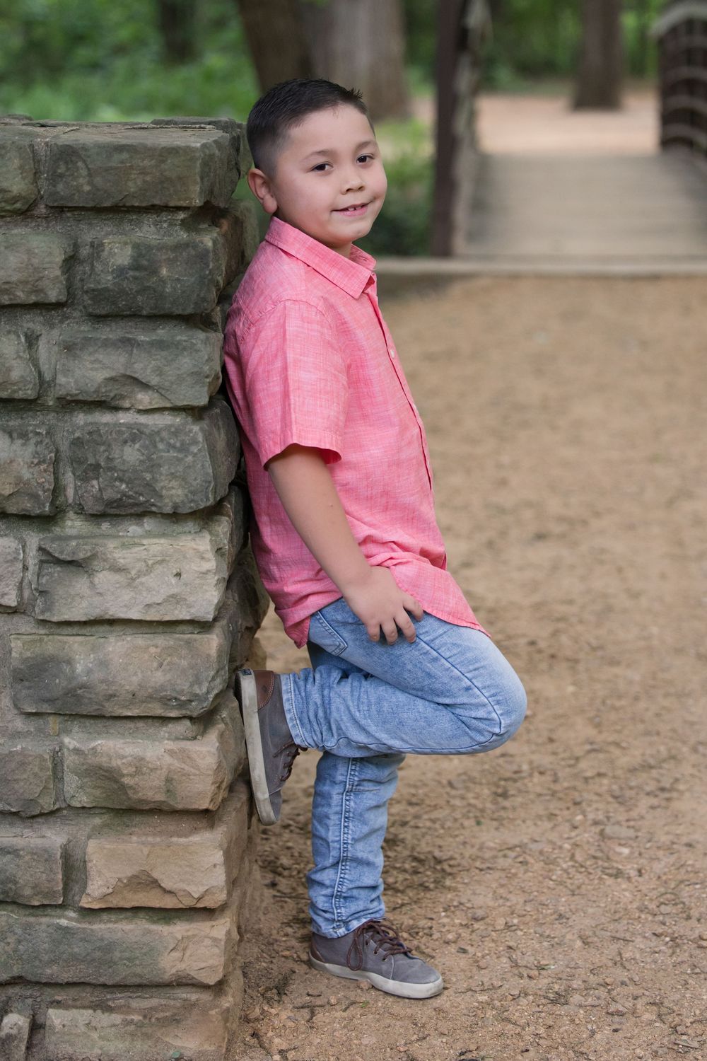 A young child in a pink shirt and jeans leans casually against a stone pillar on a dirt path through a park.