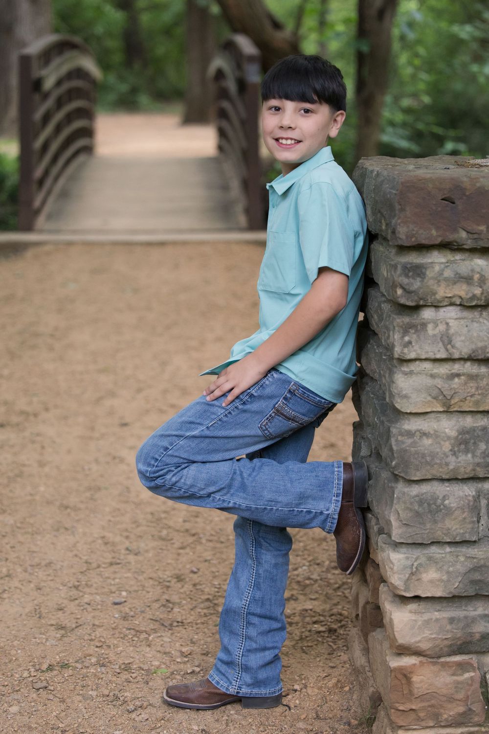Young figure in light blue shirt and jeans leans against stone pillar on dirt path with wooden bridge in background.