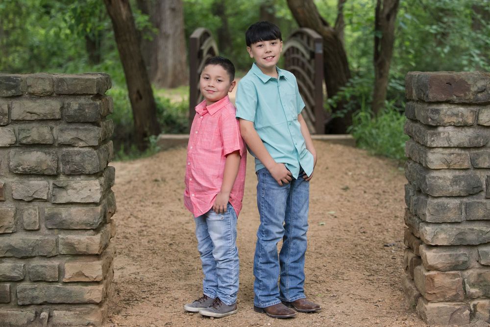 Two children in casual button-down shirts and jeans stand between stone pillars on a dirt path in a wooded area.