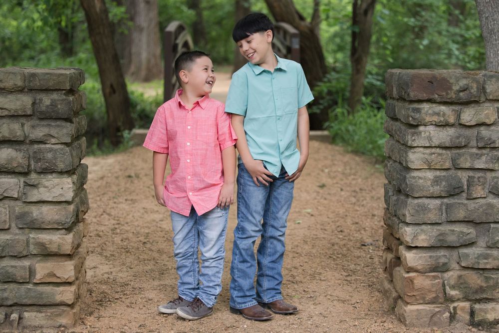 Two children wearing button-down shirts and jeans stand together on a path between stone pillars in a wooded area.