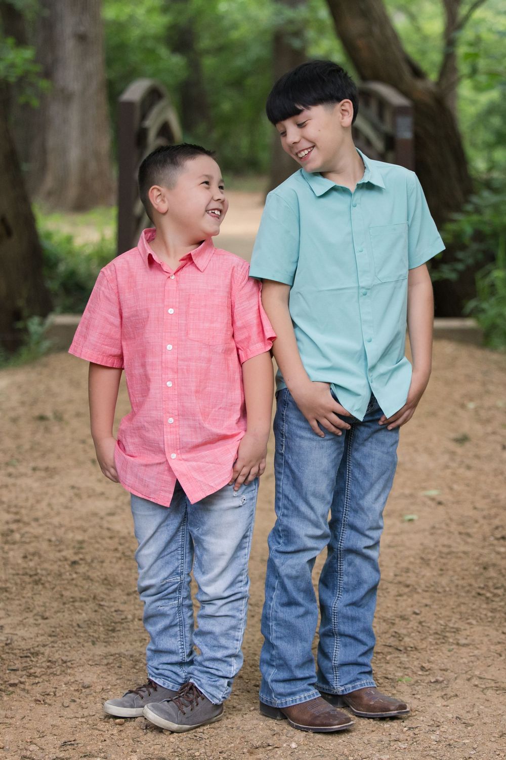 Two children wearing casual button-down shirts and jeans share a joyful moment together on a wooded path.