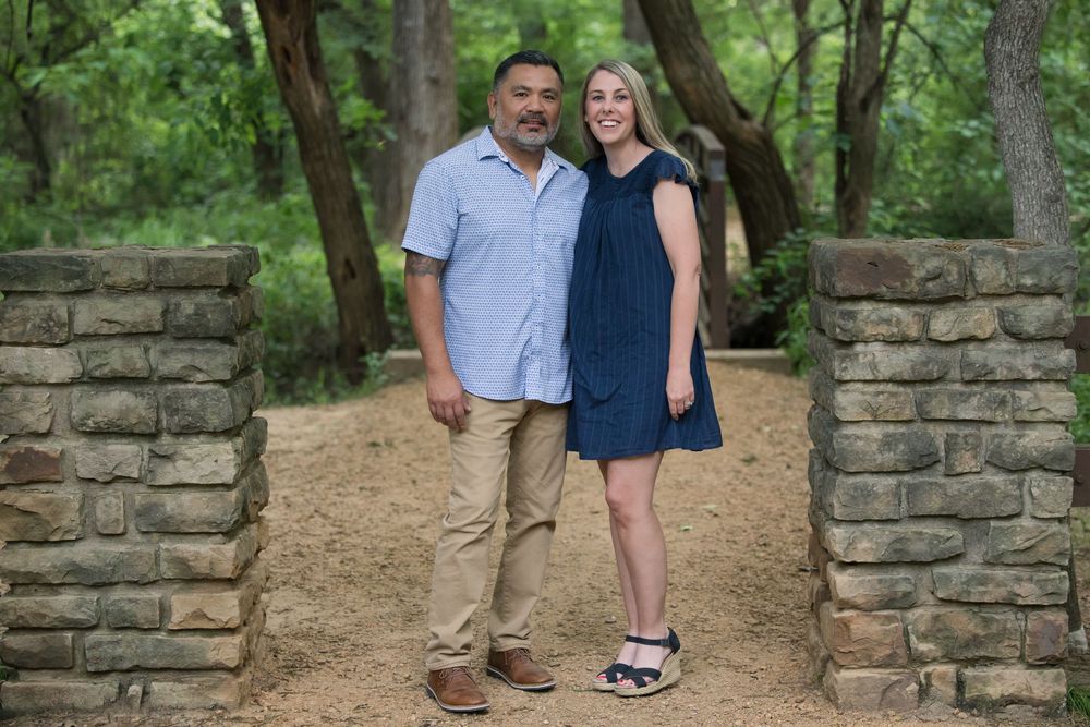 A happy couple stands together on a stone-lined path in a green wooded setting during daytime.