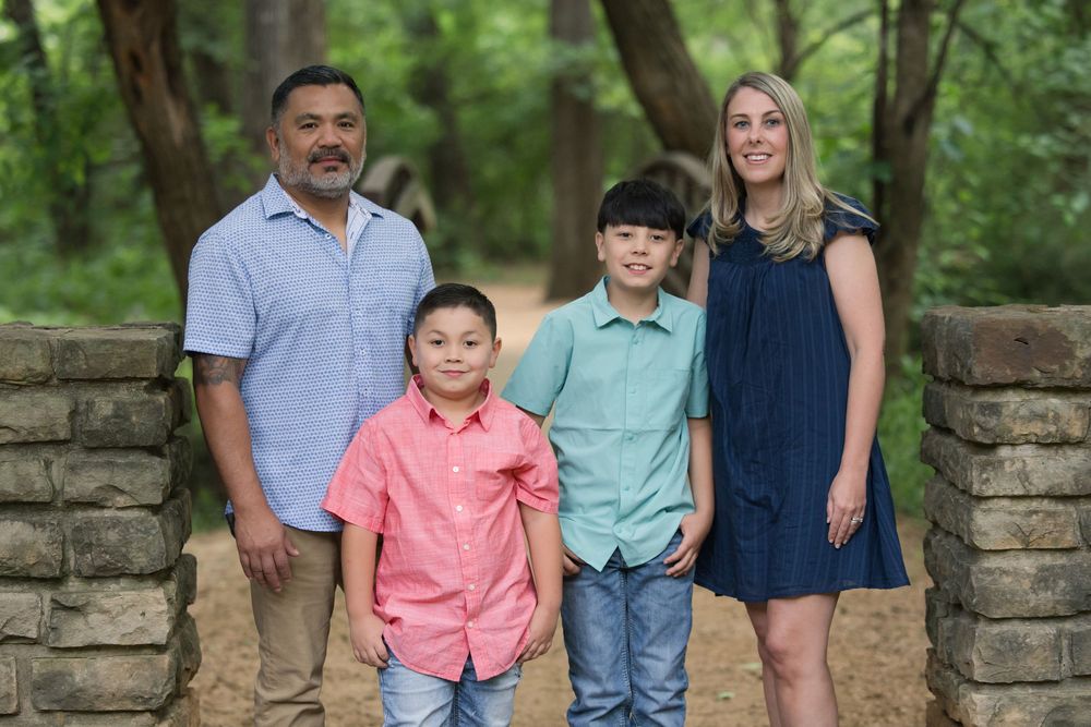 A family of four poses together in front of a stone wall surrounded by lush green trees in a park setting.