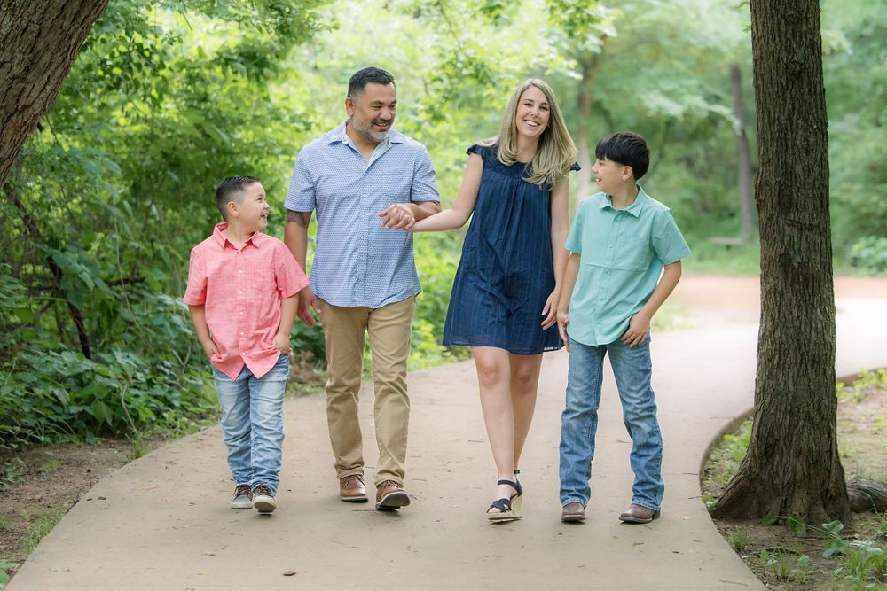 A family of four walks together smiling on a tree-lined path during a sunny summer day.