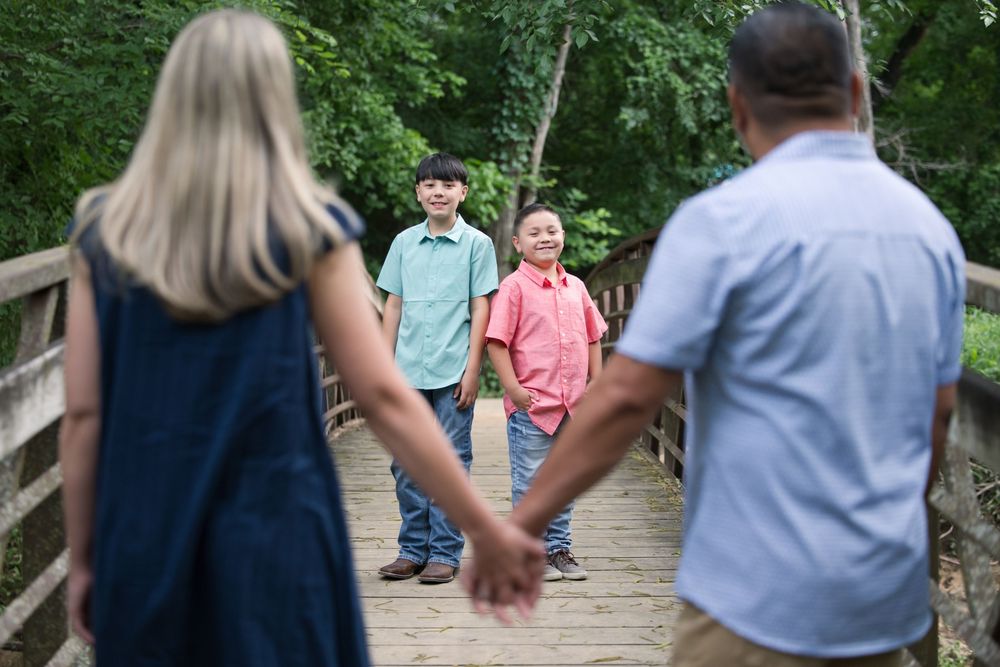 Two couples hold hands while standing facing each other on a wooden footbridge in a lush forest setting.