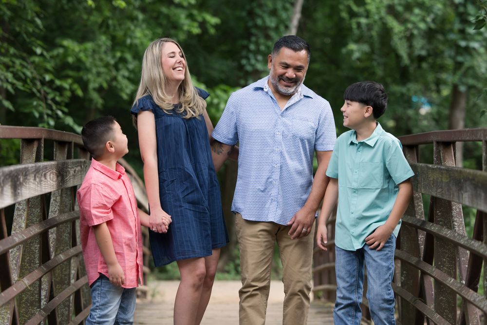 A family of four shares a joyful moment while standing together on a wooden bridge surrounded by lush greenery.