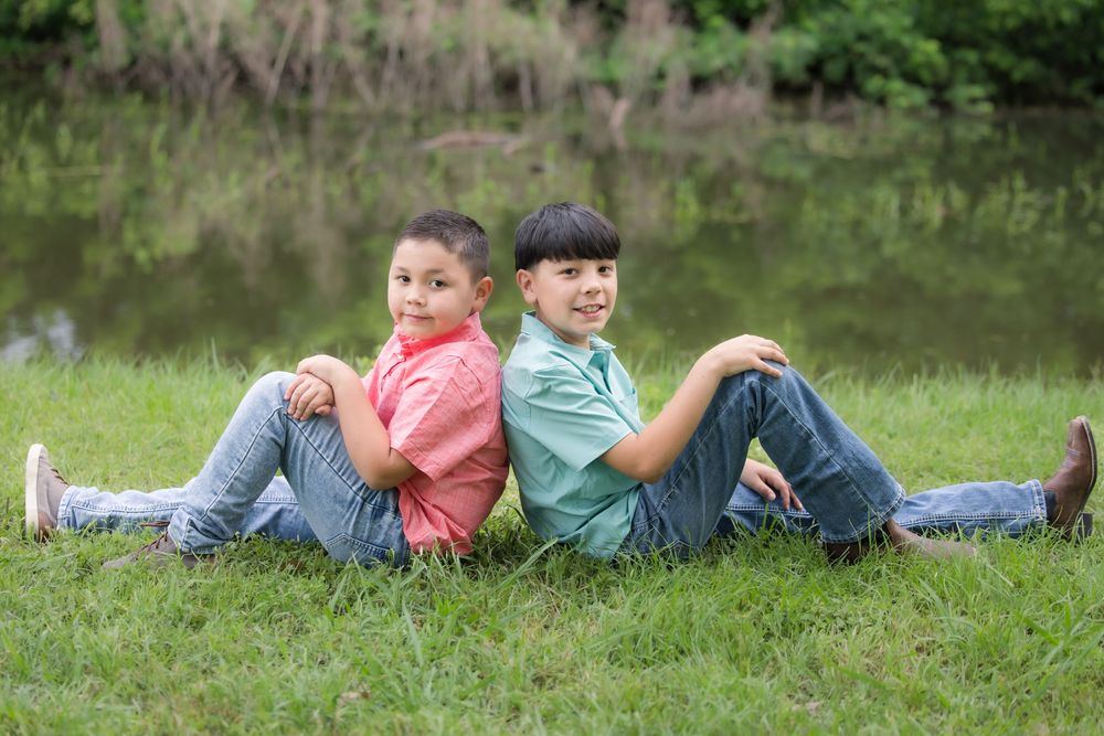 Two children sit back-to-back on grass near a pond wearing casual clothing and smiling on a sunny day.
