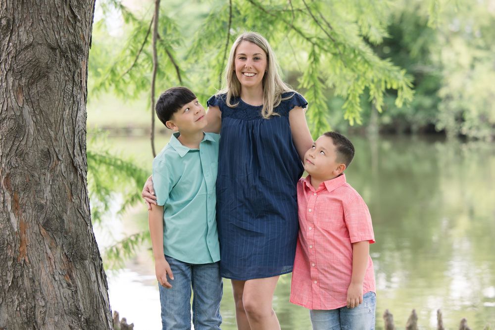 A family stands together smiling by a tree next to a peaceful pond on a sunny summer day.