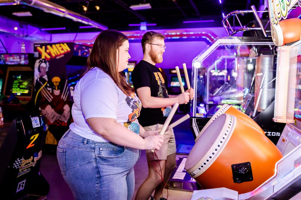 Two people playing arcade games in a dimly lit retro arcade with neon purple lighting and gaming cabinets.