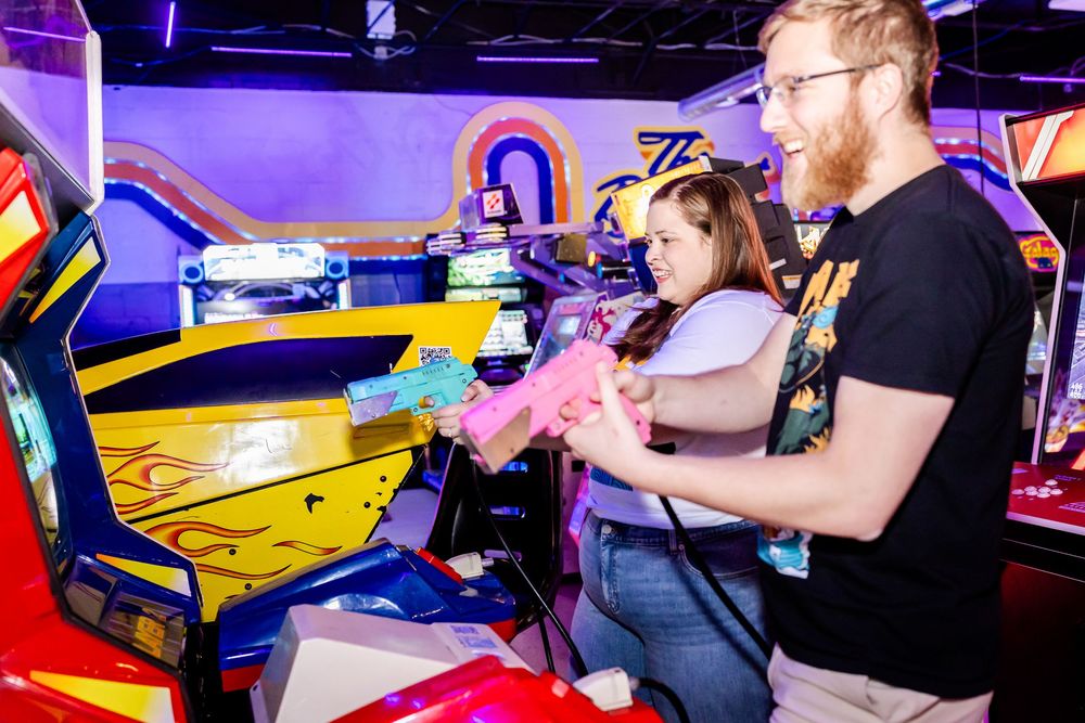 Two people playing a colorful arcade racing game together in a vibrant neon-lit gaming venue.