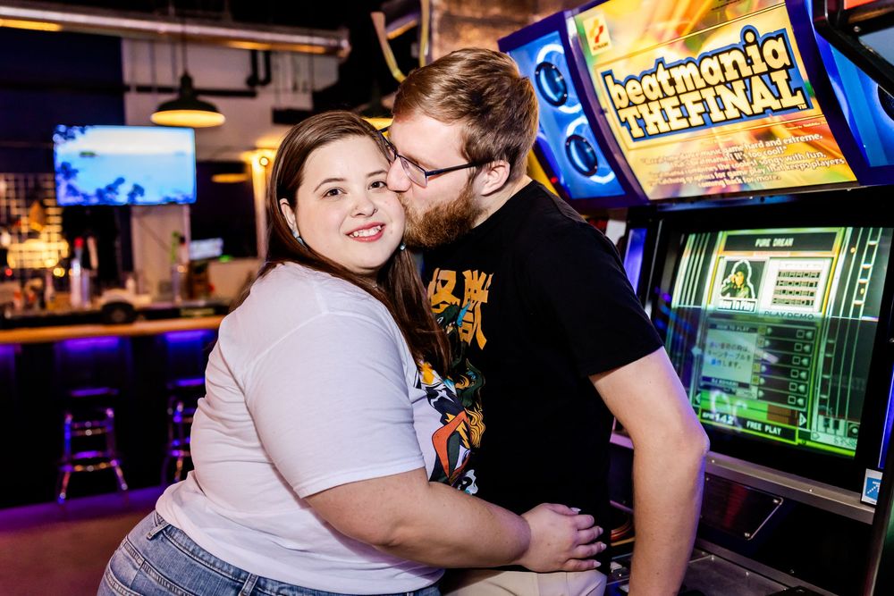 A couple shares a tender moment while sitting at an arcade bar surrounded by glowing game cabinets and screens.