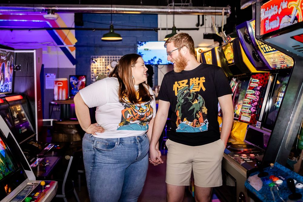 Two arcade enthusiasts interact while playing games in a dimly lit retro arcade setting.