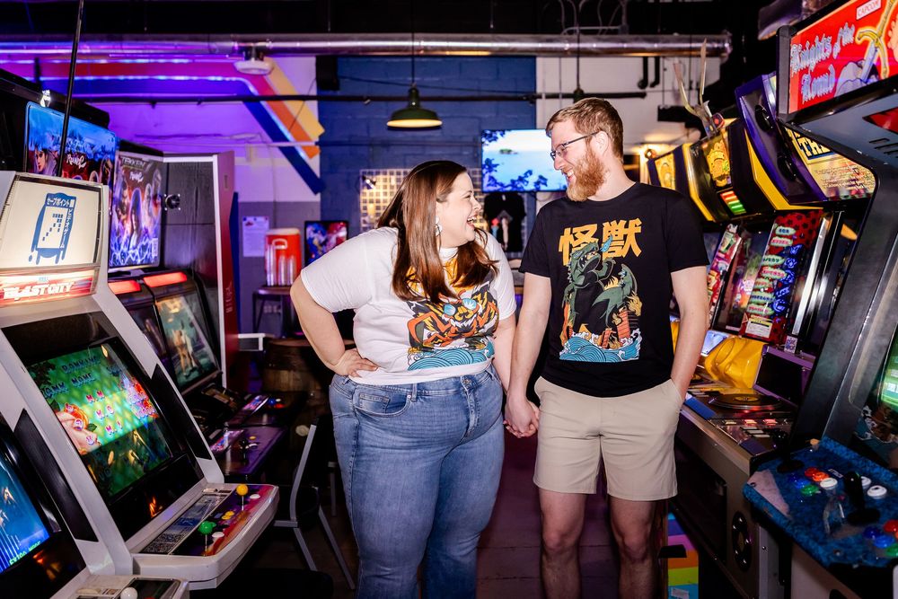Two people play arcade games together in a retro arcade room with classic cabinets and purple neon lighting.
