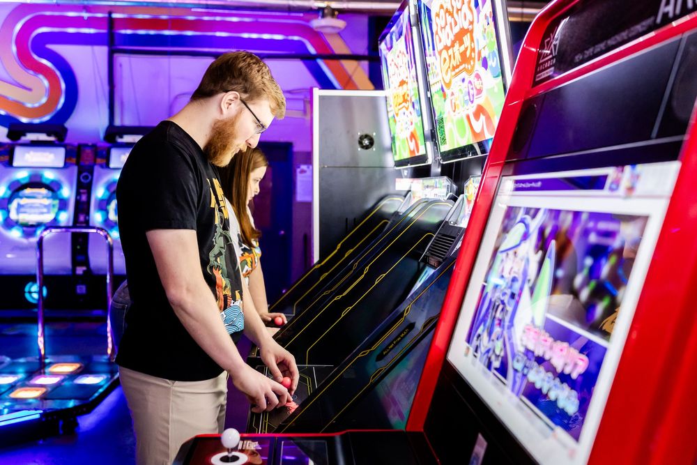 Someone wearing a black t-shirt plays an arcade game with neon purple lighting in a classic arcade setting.
