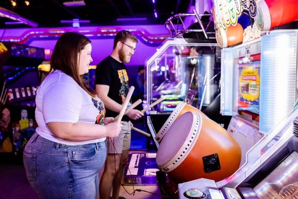 Two people enjoy playing arcade games in a colorful arcade with purple lighting and drum-based rhythm game.
