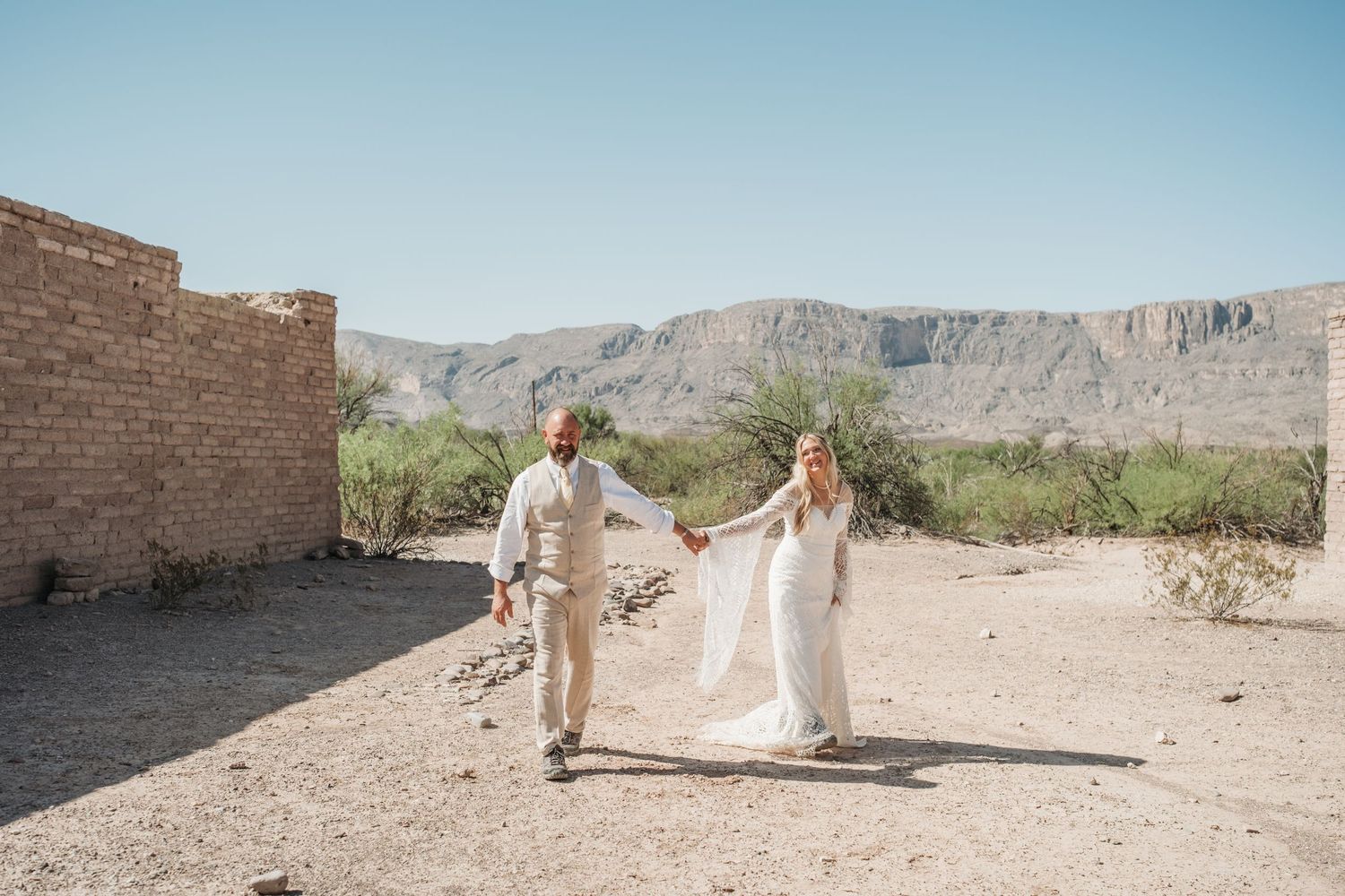 A desert wedding scene unfolds near adobe ruins with mountain backdrop.
