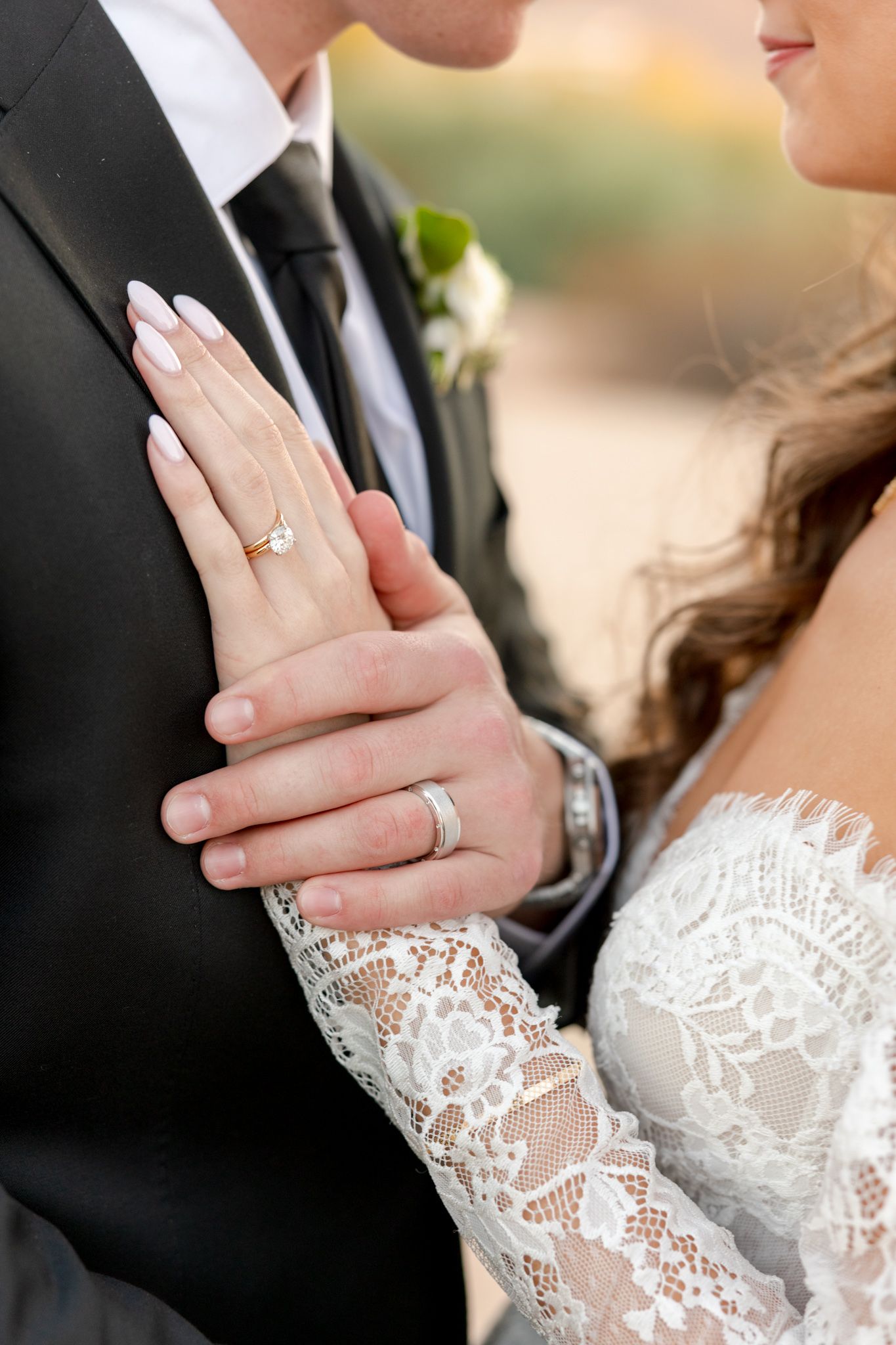 A close-up wedding photo showing hands with rings and delicate lace sleeves against formal attire.