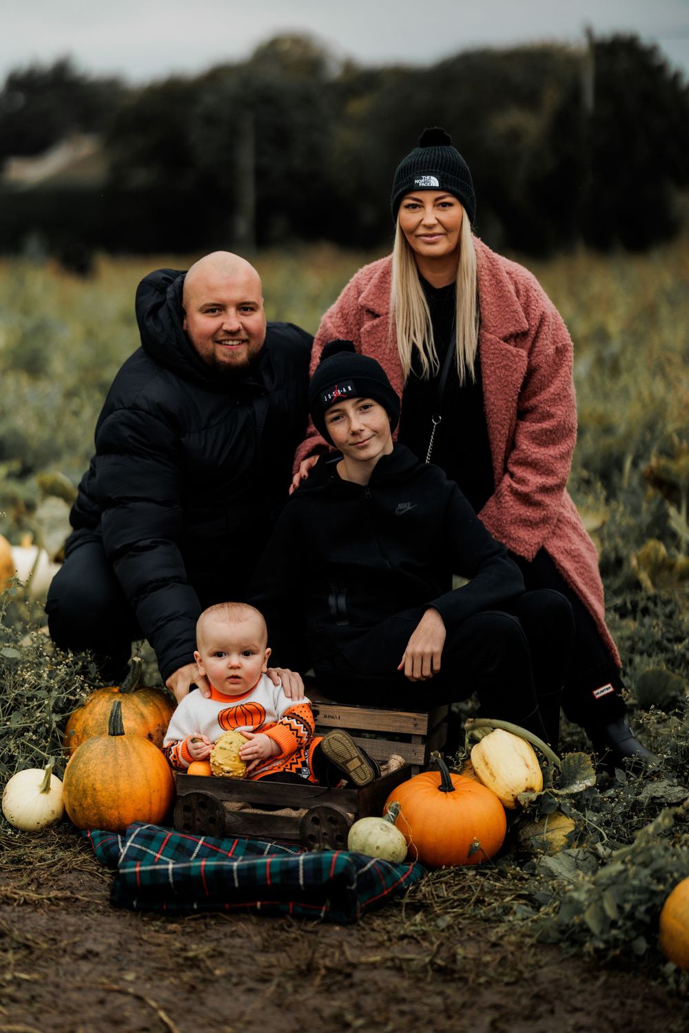 A family group poses with pumpkins and gourds on a plaid blanket in a field.
