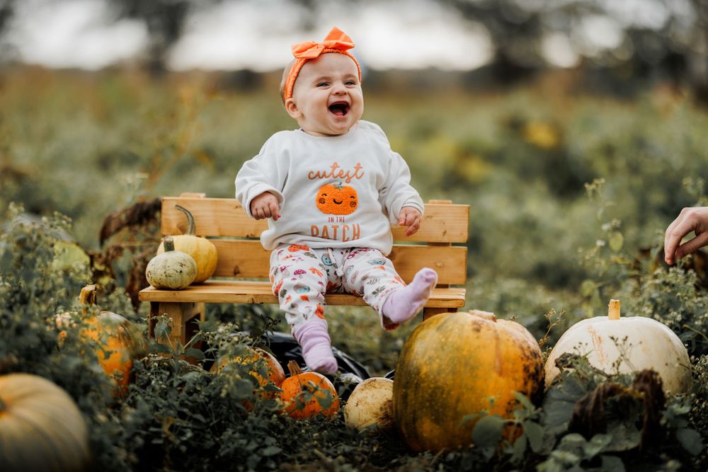 Happy baby in pumpkin-themed outfit sits among harvest gourds in outdoor setting.