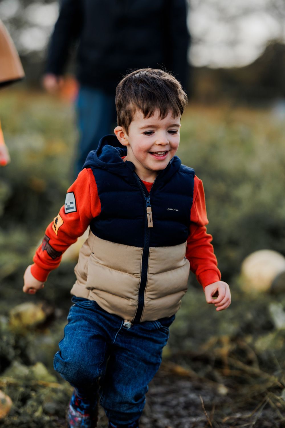 A child in a puffy vest and orange sleeves runs through a pumpkin patch.