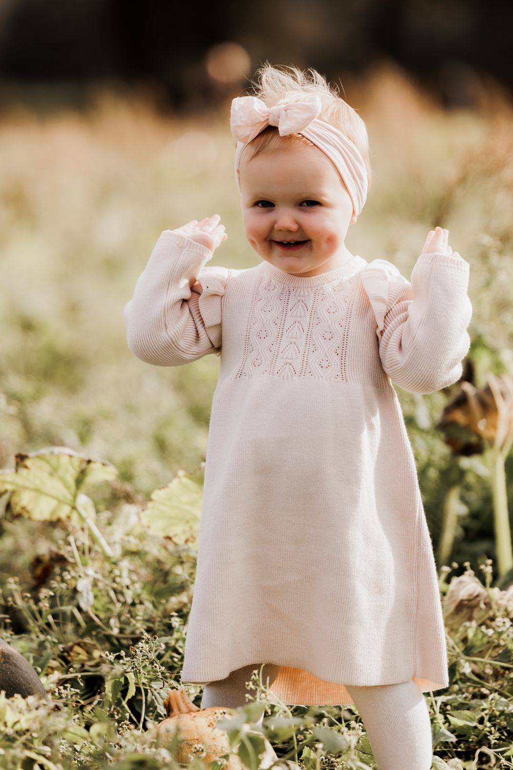 Baby in white dress and bow headband smiling joyfully in outdoor setting.