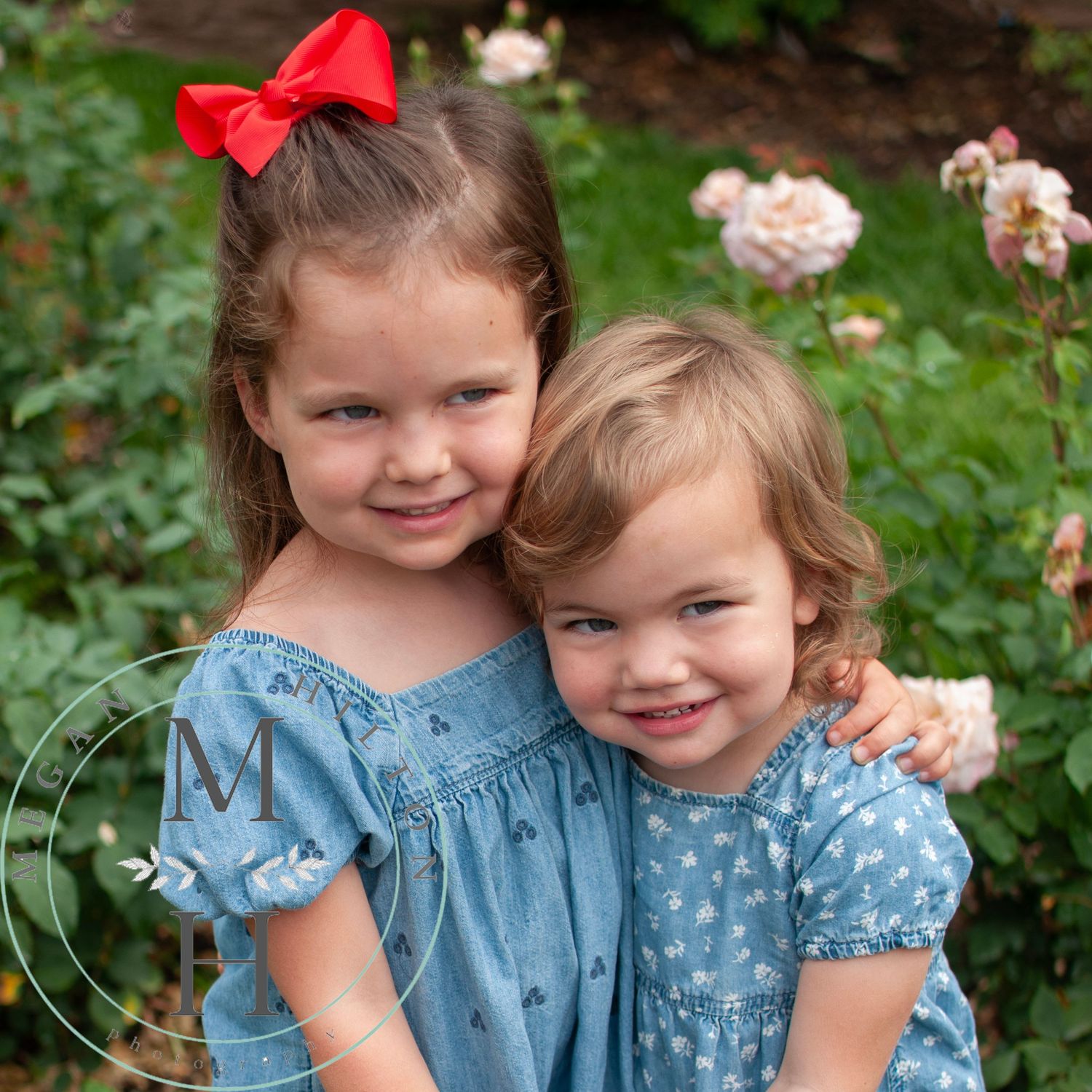Two young children wearing matching blue floral dresses pose together in a garden with pink roses in the background.