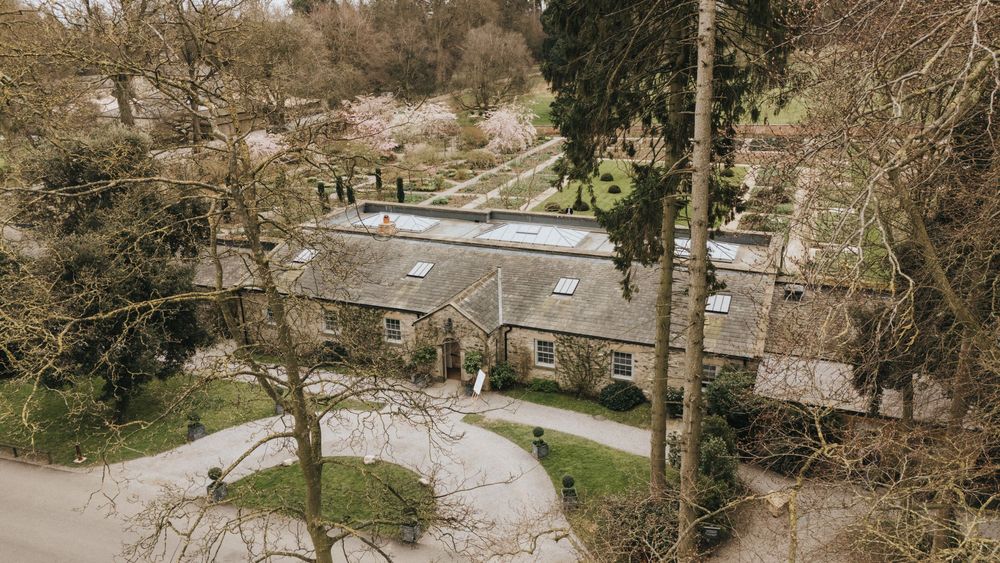 Aerial view of a rustic stone cottage with a curved driveway nestled among tall trees in a rural setting.