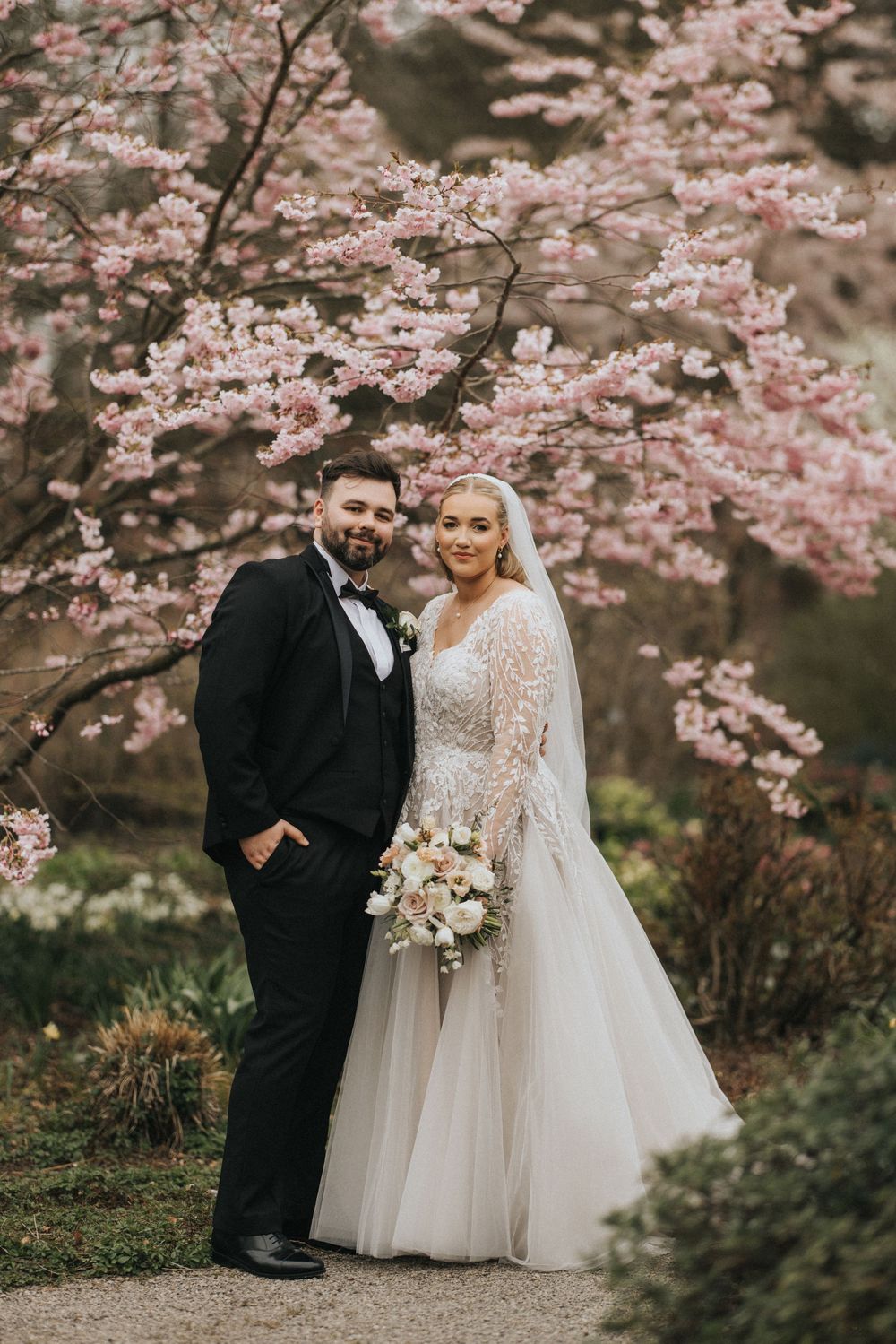 A romantic wedding portrait under blooming pink cherry blossoms during spring with a white floral bouquet.