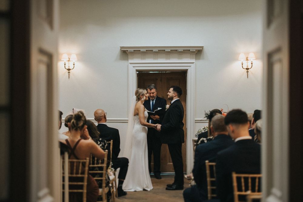A modern wedding ceremony takes place in a bright white room with wooden chiavari chairs and wall sconces.