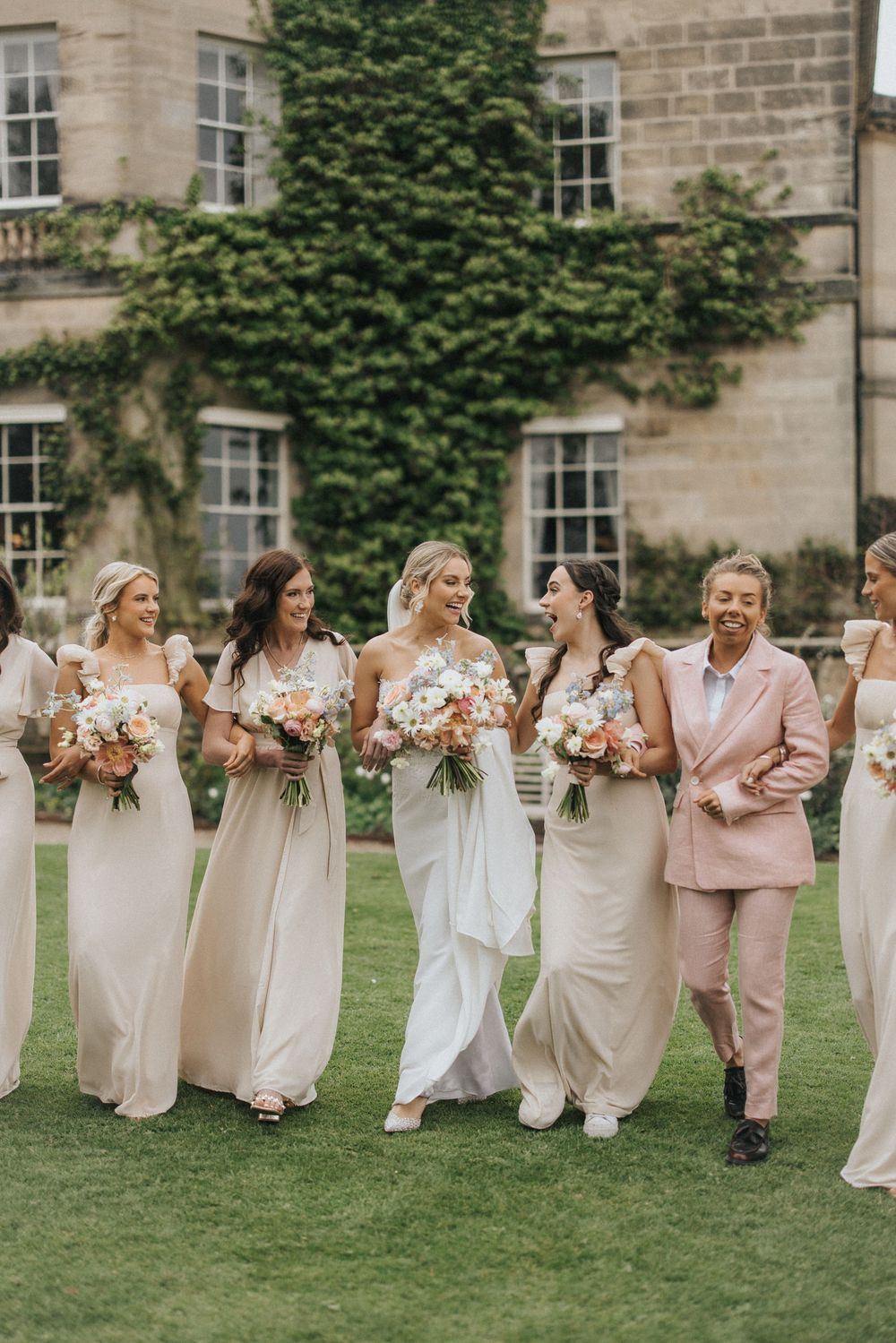 Wedding party in white and blush pink attire poses together on a grassy lawn in front of historic stone building.