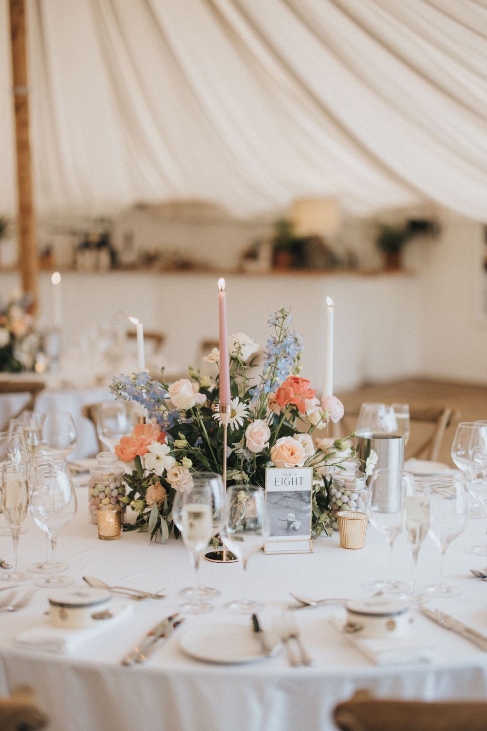 Elegant wedding reception table setting with white candles, peach and blue floral centerpiece in a white tent venue.