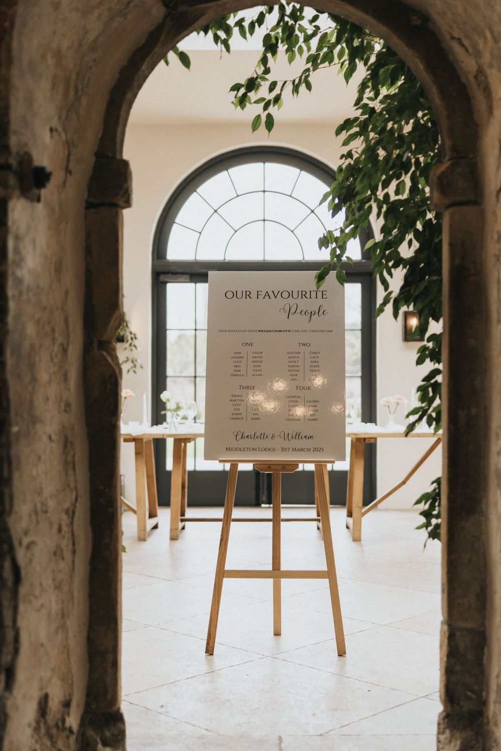 A wedding playlist sign on a wooden easel stands framed by an arched doorway with green foliage and natural lighting.