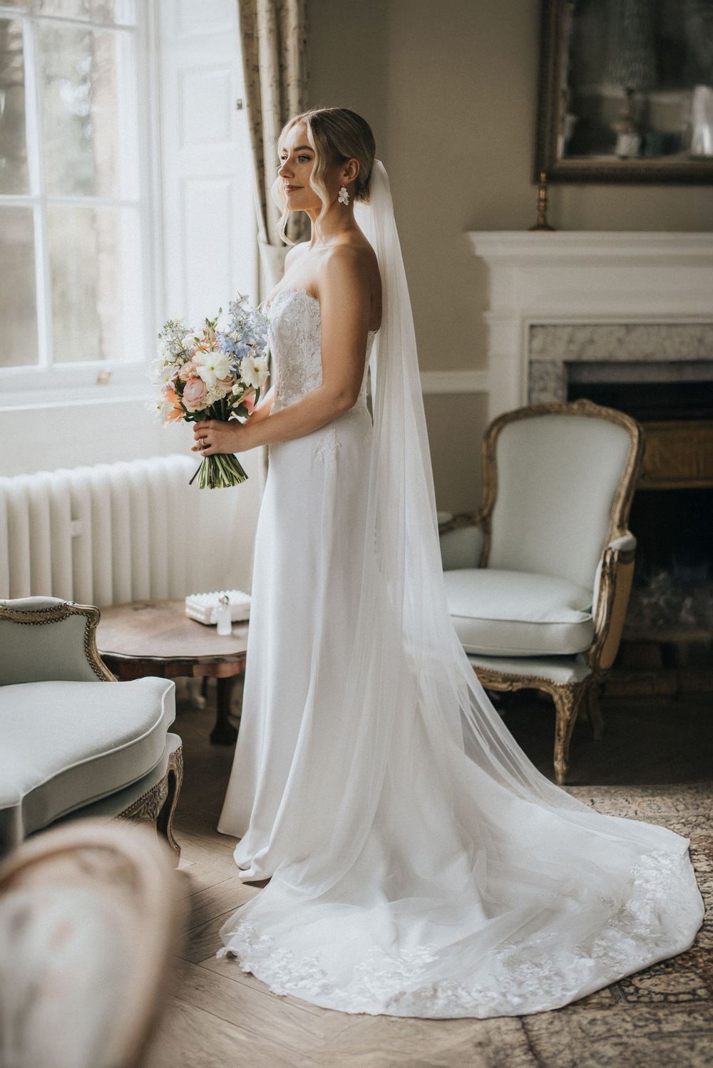 A bride in a classic white wedding gown with a flowing veil stands elegantly in a bright, vintage-styled interior space.