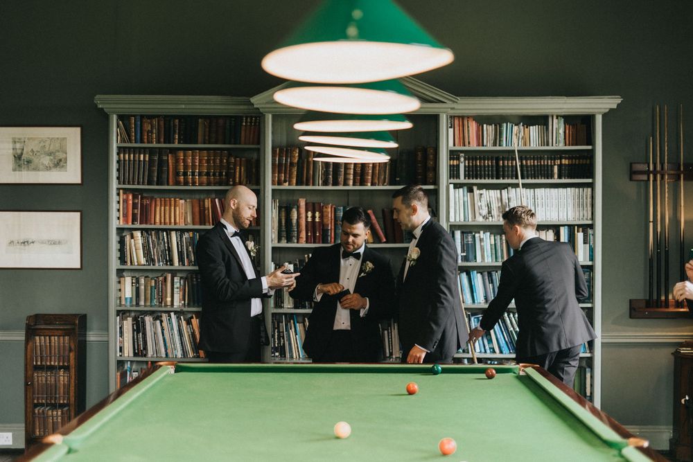 Well-dressed people gather around a pool table in a stylish library room with bookshelves and a green hanging lamp.
