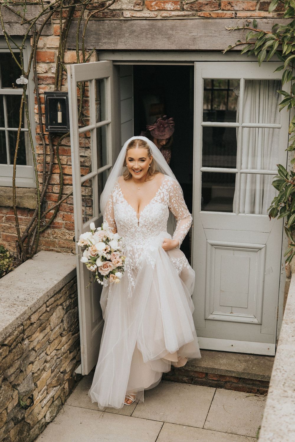 Beautiful bride in long sleeve lace wedding dress steps out of rustic doorway holding blush and white floral bouquet.