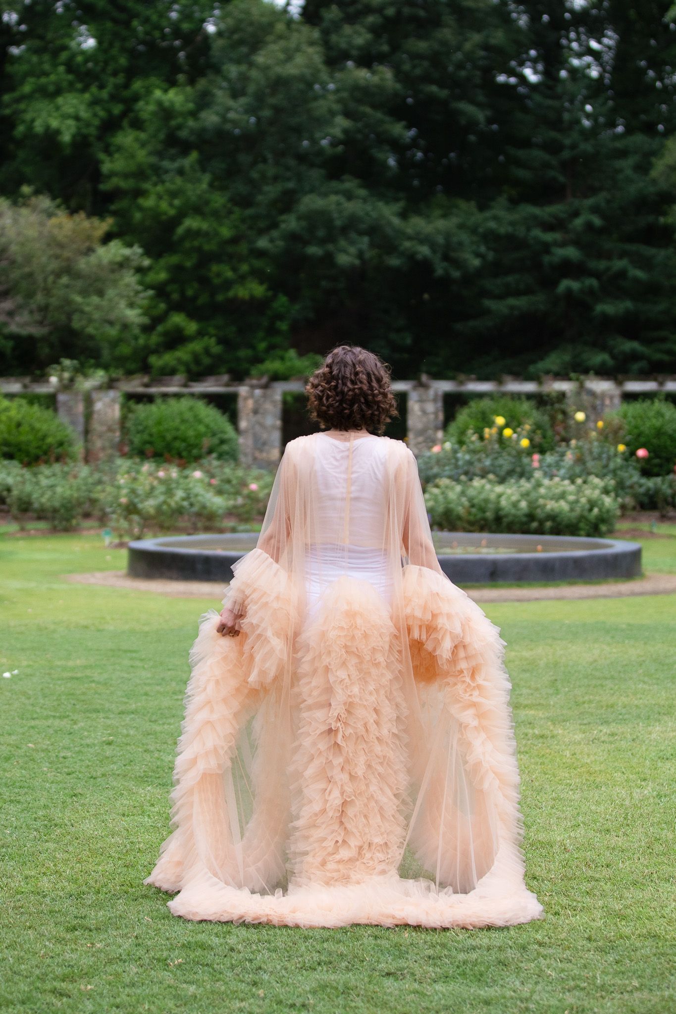 A woman in a flowing peach-colored tulle gown with ruffles poses outdoors at the Raleigh rose Garden.