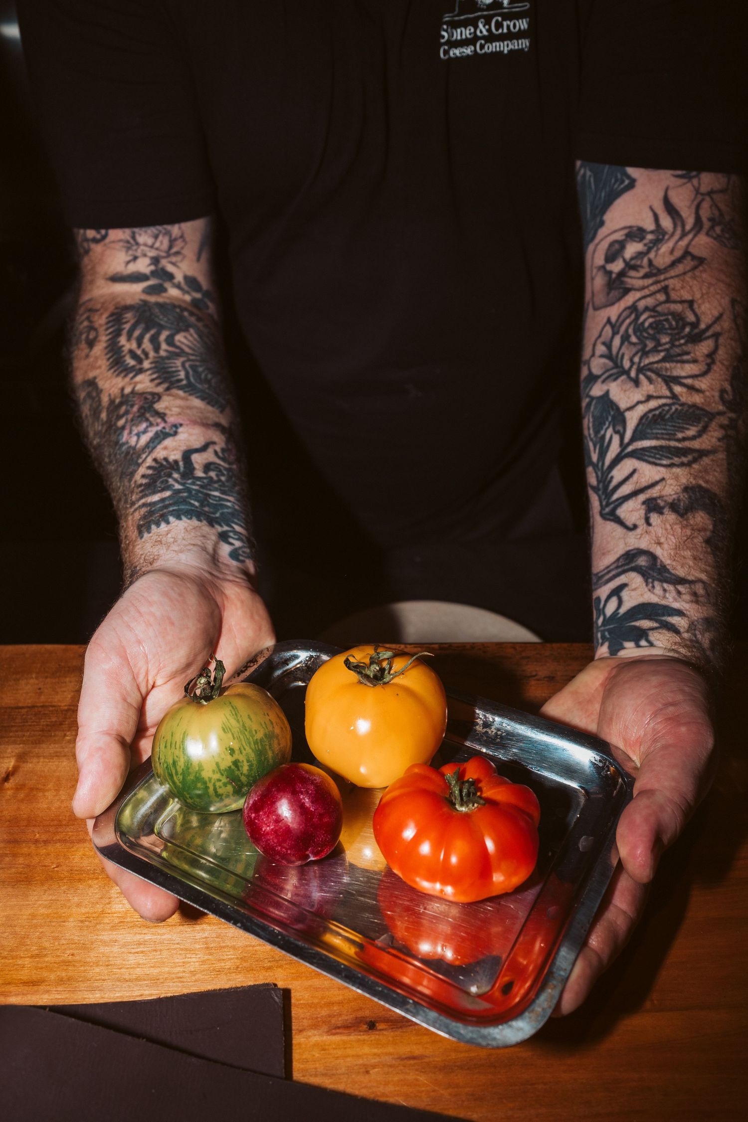 Tattooed arms holding a metal tray with vibrant fresh produce, including heirloom tomatoes and a radish, on a wooden table