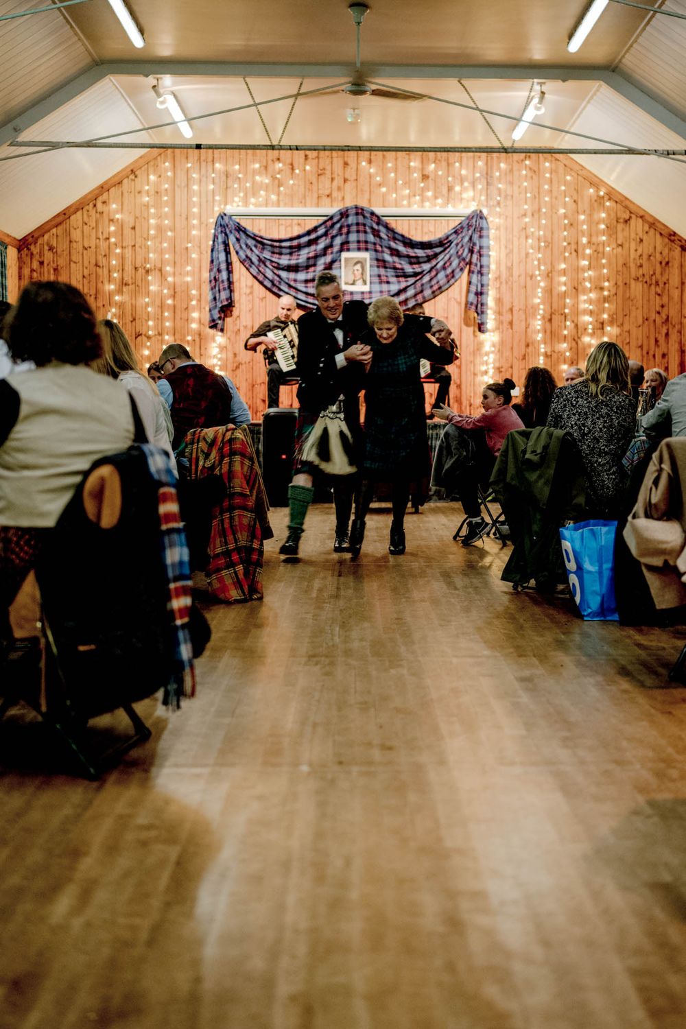 People dancing and celebrating at an indoor event venue decorated with string lights and purple drapes.