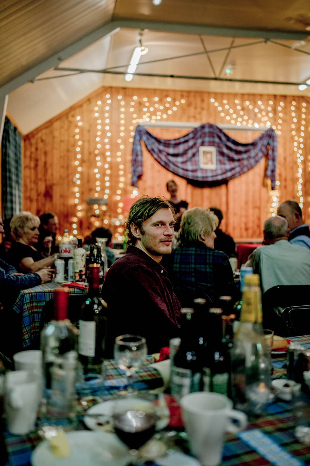 A festive indoor dinner party with string lights and blue drapes decorating a wooden wall in the background.