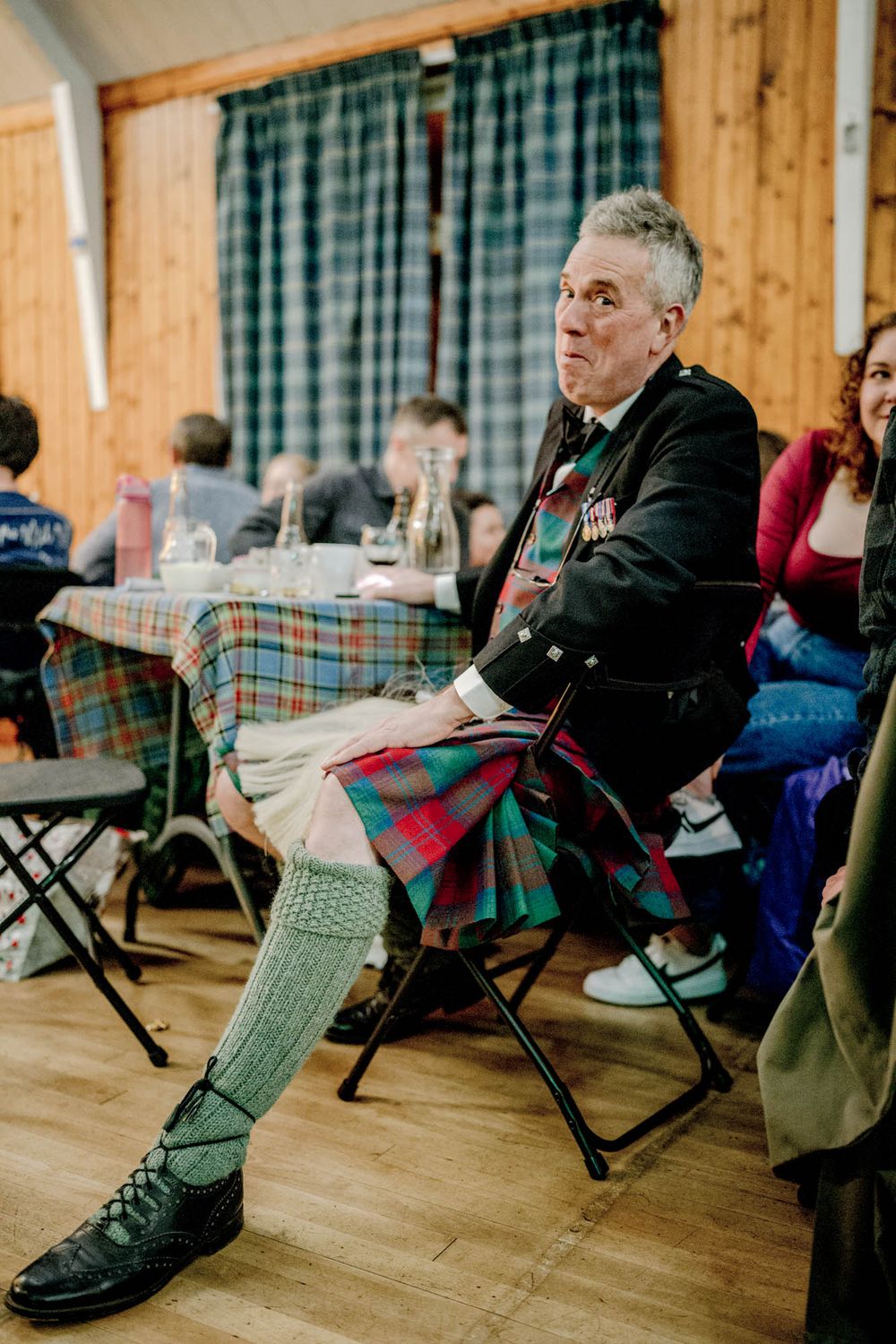 Traditional Scottish attire featuring a tartan kilt and green knee socks at an indoor gathering with plaid curtains.
