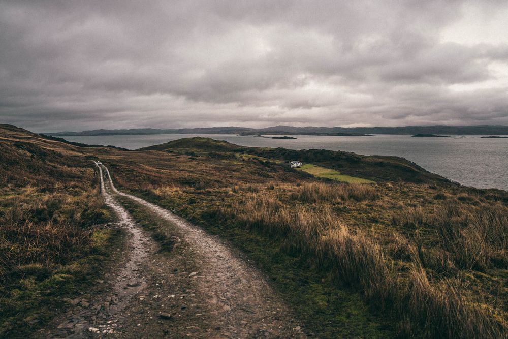 A winding dirt path leads along rugged coastal cliffs with moody overcast skies and ocean views in the distance.