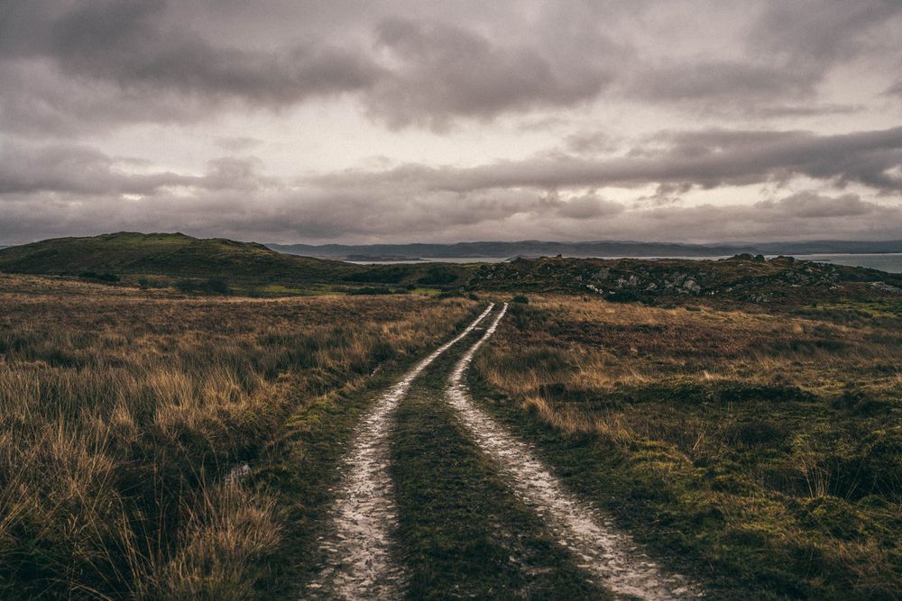 A winding dirt path leads through moody moorland under dark stormy clouds in the Scottish highlands.