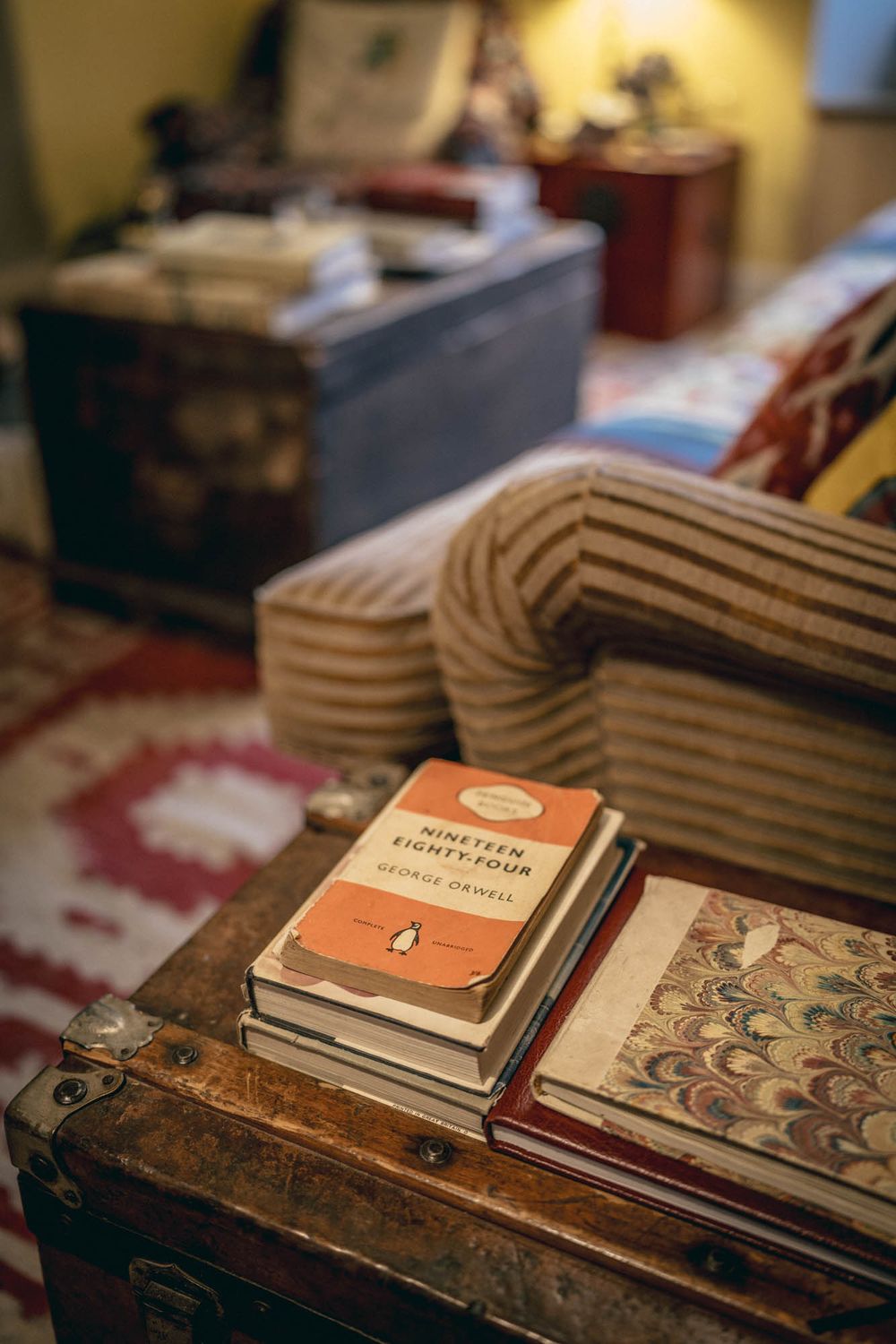 A vintage orange Penguin book sits atop a stack of books on an old wooden trunk with cushions in the background.