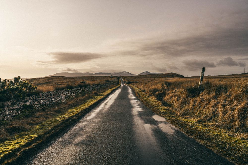A winding country road stretches through green rolling hills under dramatic cloudy skies during sunset.