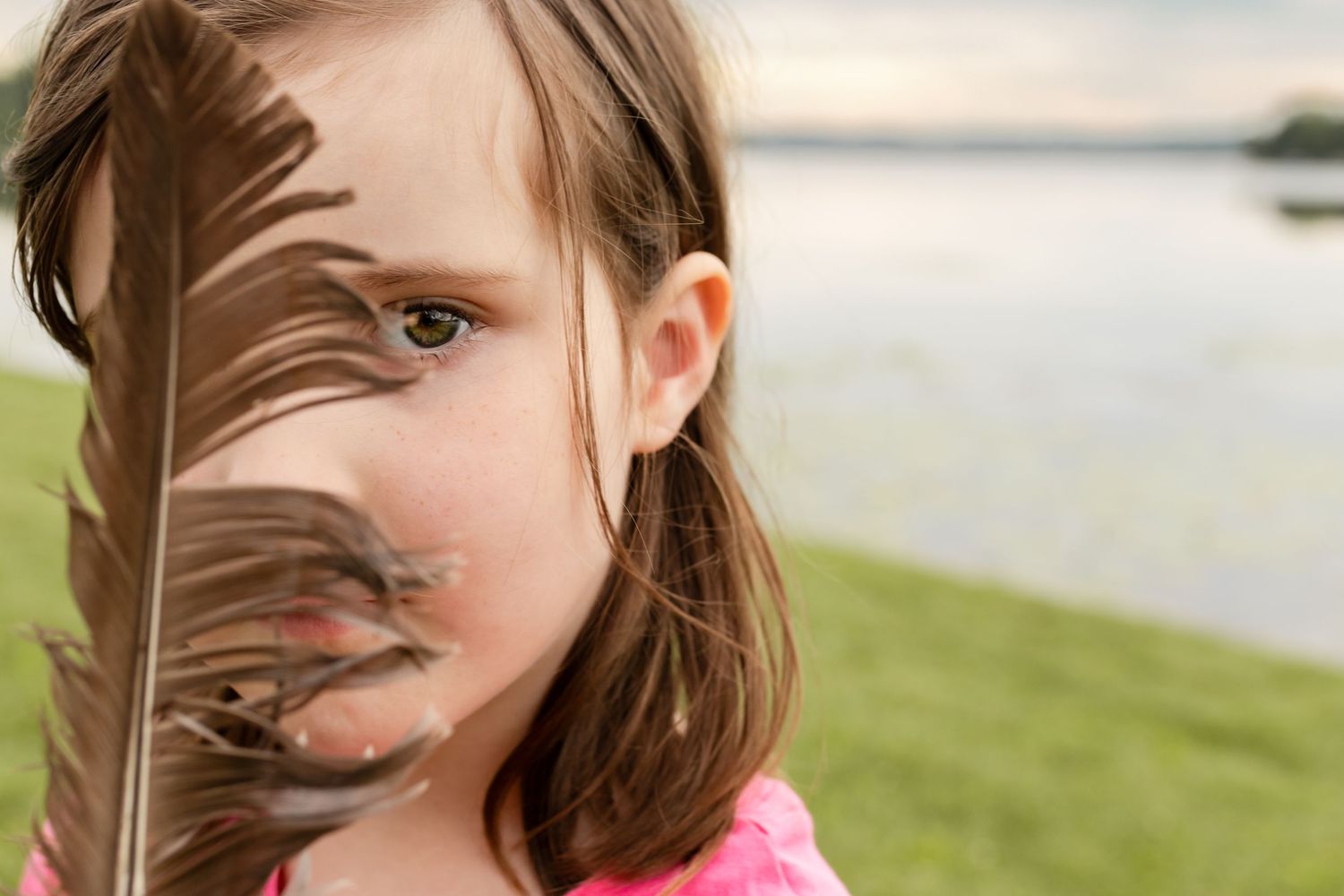 A young girl in pink top peaks through a gap in a feather at the camera, green grass and a calm lake behind her.