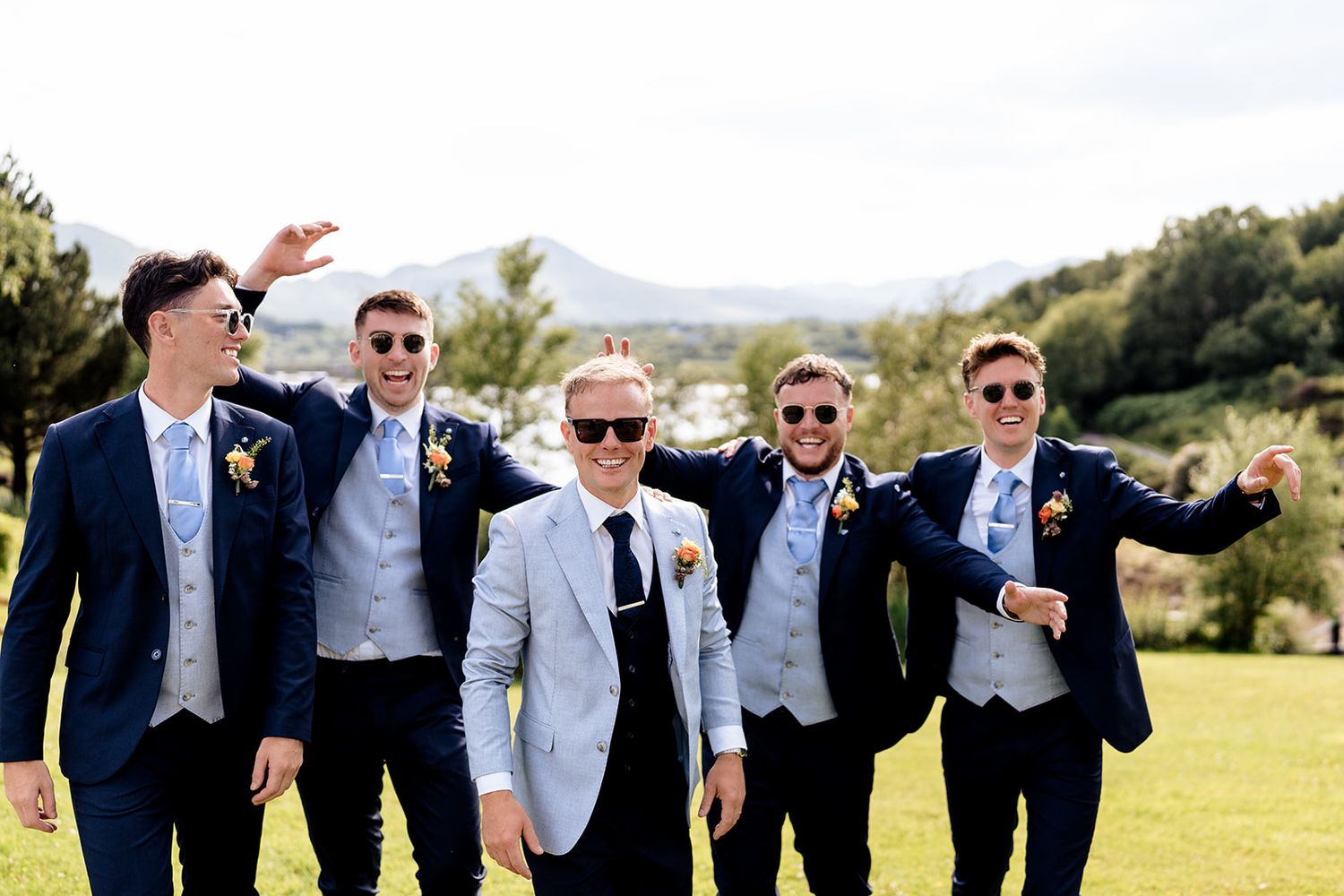 A group of groomsmen in matching black suits and sunglasses pose playfully on a sunny hillside.