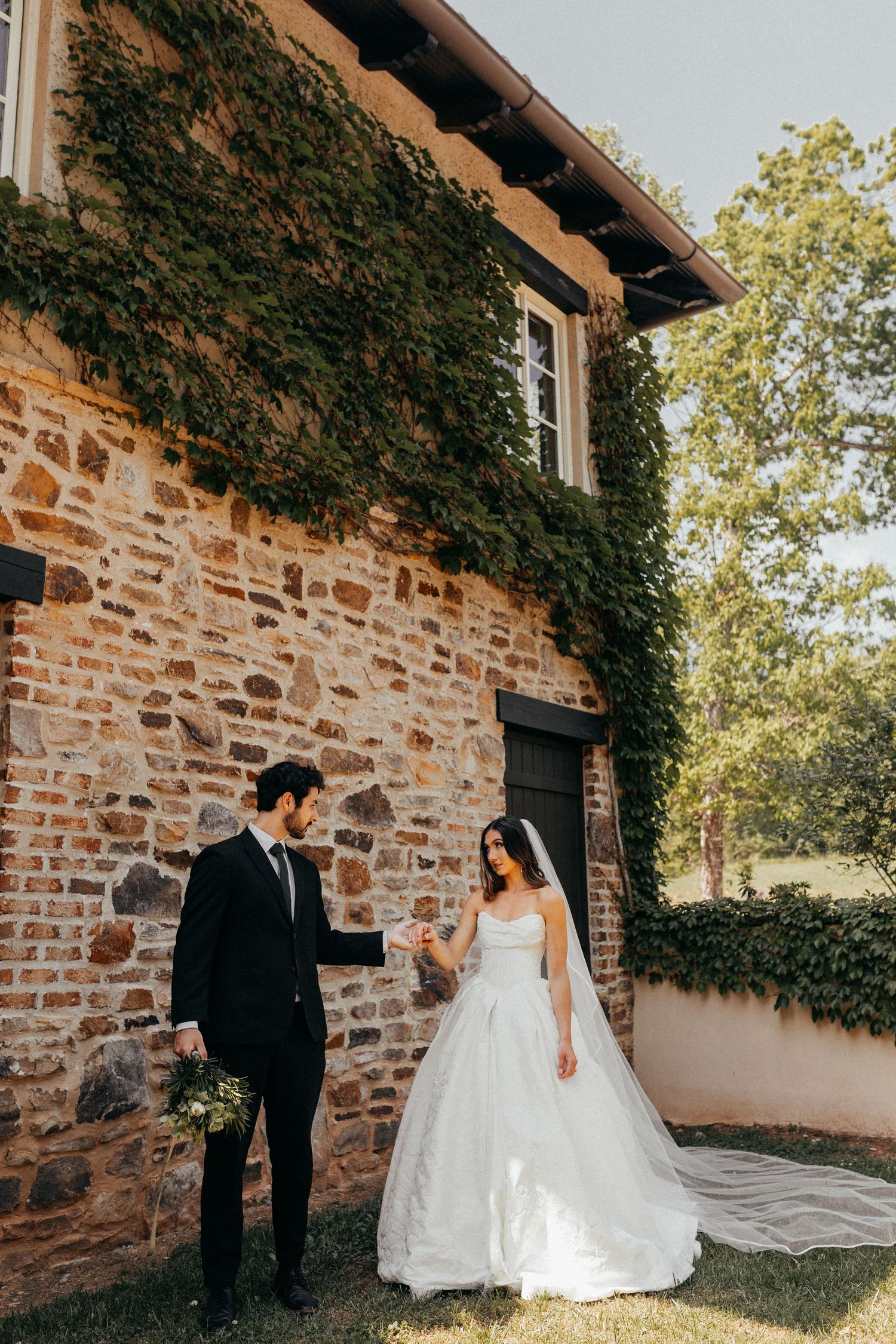 A romantic moment captured outside a rustic stone building covered in ivy on a sunny wedding day.