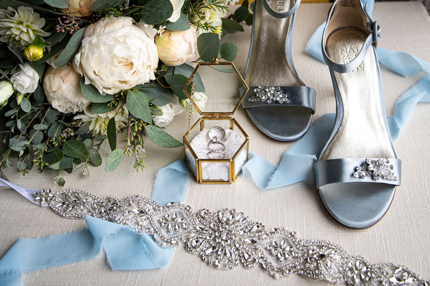 Wedding accessories including blue shoes, floral arrangements, and jewelry on a light gray surface.