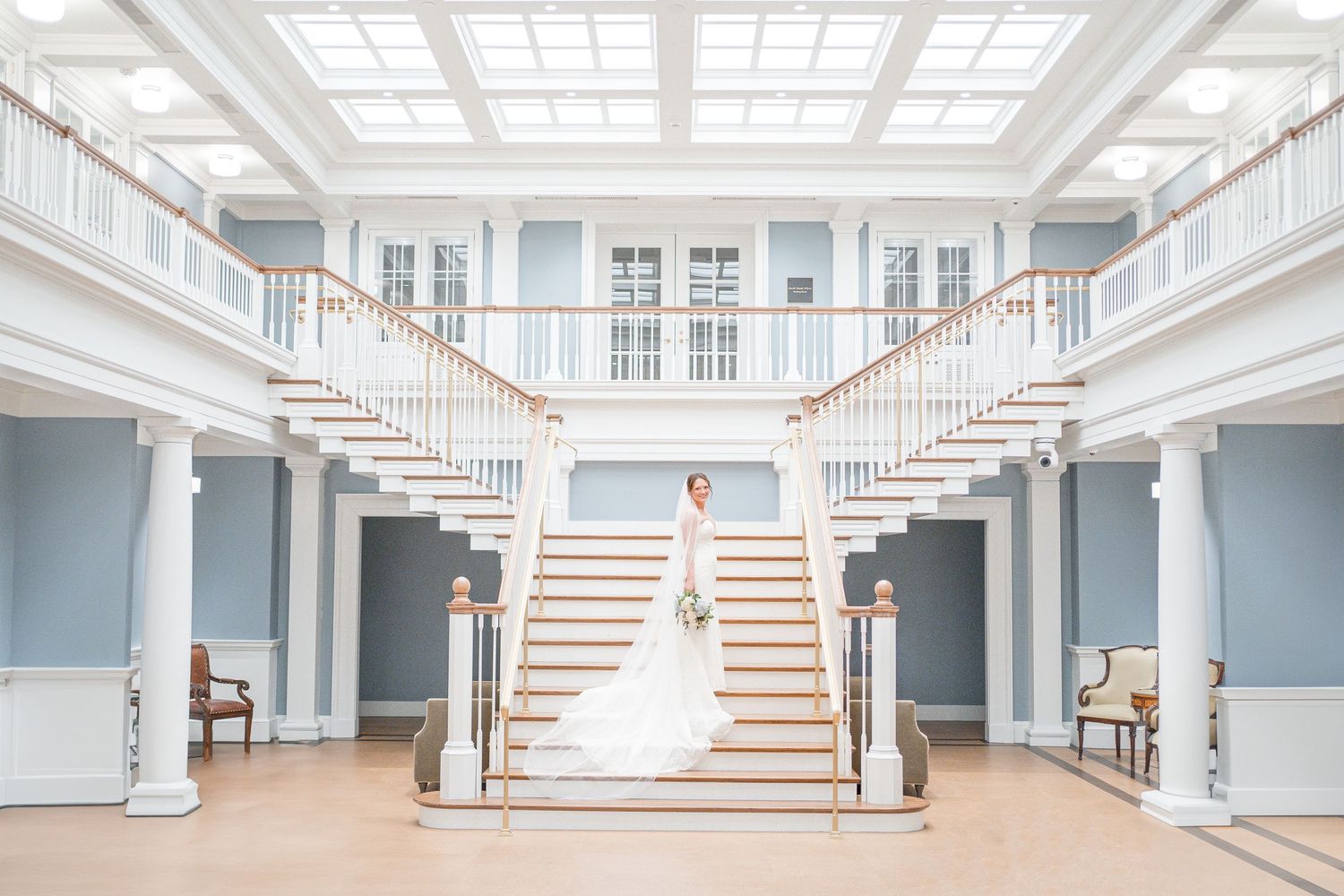 A person in a wedding gown stands centered on an elegant grand staircase in a light blue and white classical interior.